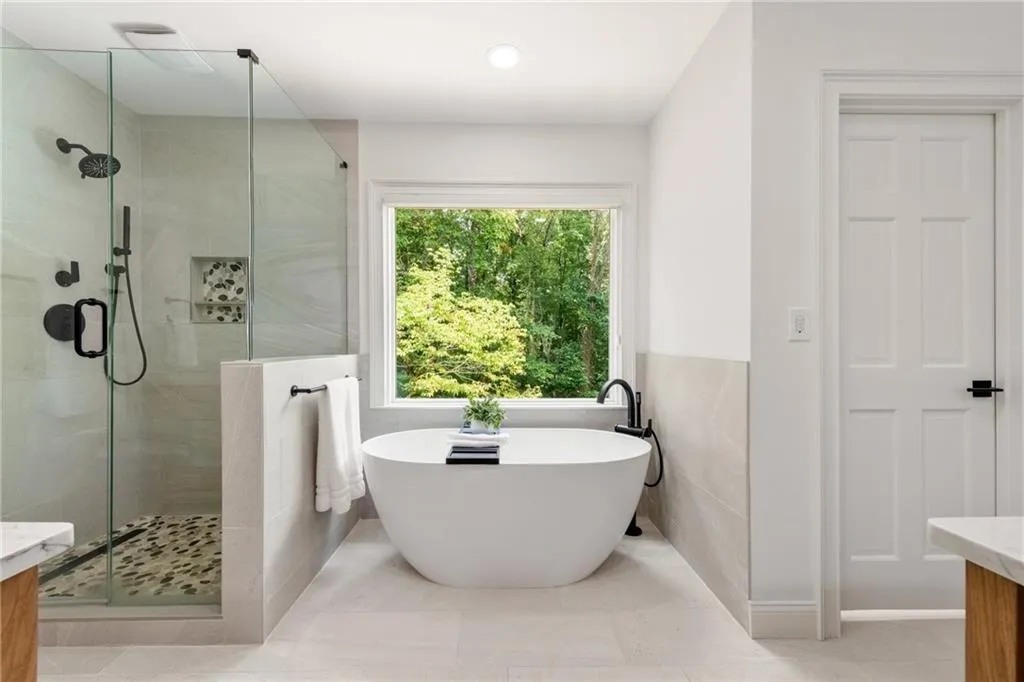 The spa-inspired primary bath features a large soaking tub framed by a picture window that provides both privacy and natural sunlight. A separate glass-enclosed shower with a pebble floor adds a touch of luxury, while porcelain tile flows seamlessly throughout the space for a sleek, contemporary finish.