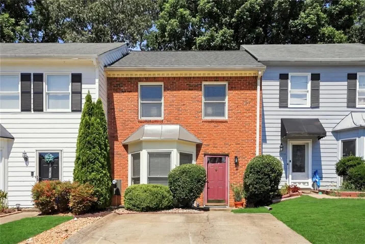 Brick facade townhome with a red entry door, bay window, and mature landscaping