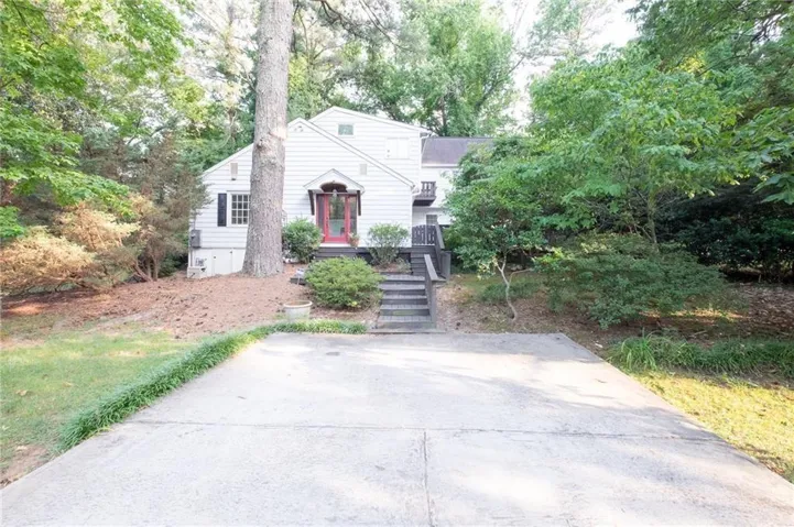 Charming cottage nestled on a wooded lot featuring a concrete driveway, front entry with decorative wreath, horizontal siding exterior, and mature trees providing natural screening