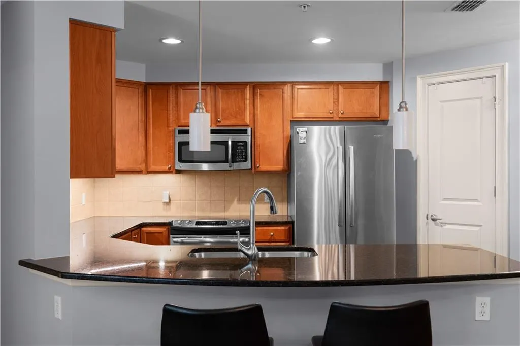 Kitchen featuring stainless steel appliances, a peninsula, dark stone countertops, wood finish cabinetry, and pendant lighting