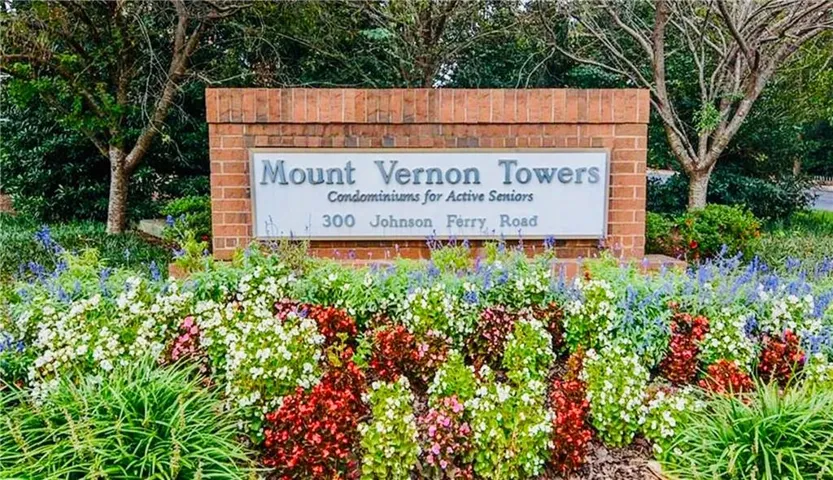 Brick monument sign with an embedded plaque, framed by diverse flowering plants and established trees