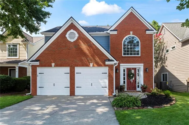 Brick facade residence featuring a two-car garage with white doors, a red entry door with sidelights, an arched window with grilles, and manicured landscaping
