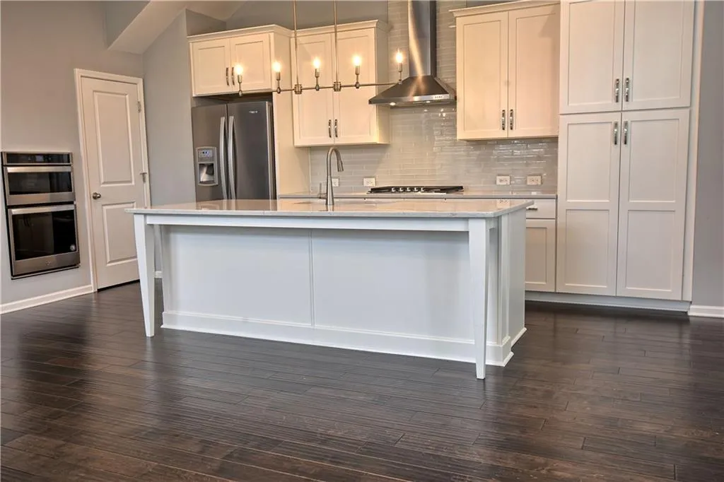 Kitchen featuring decorative backsplash, a kitchen island with sink, dark wood-style flooring, appliances with stainless steel finishes, and wall chimney exhaust hood