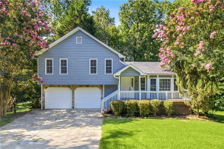 Split-level residence featuring blue siding, a two-car garage with stone accents, a covered front porch with white railing, a front lawn, and mature trees