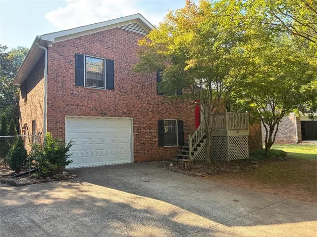 Brick exterior with white garage door, featuring an exterior staircase leading to a raised deck with lattice skirting, and mature tree landscaping