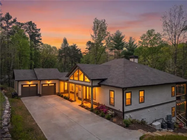 Modern farmhouse residence showcasing a two-car garage with contemporary paneling, expansive front facade with multiple large windows and glass-paneled entry, gabled roofline with stone and siding exterior, concrete driveway, and mature landscaping with evergreen and deciduous trees throughout the property