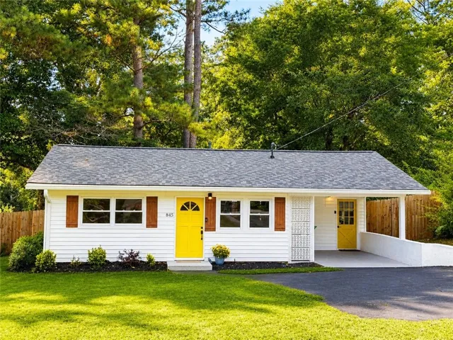 White siding exterior with a gray shingle roof, vibrant yellow entry doors, wood-tone louvered shutters, an attached carport, and a manicured front lawn