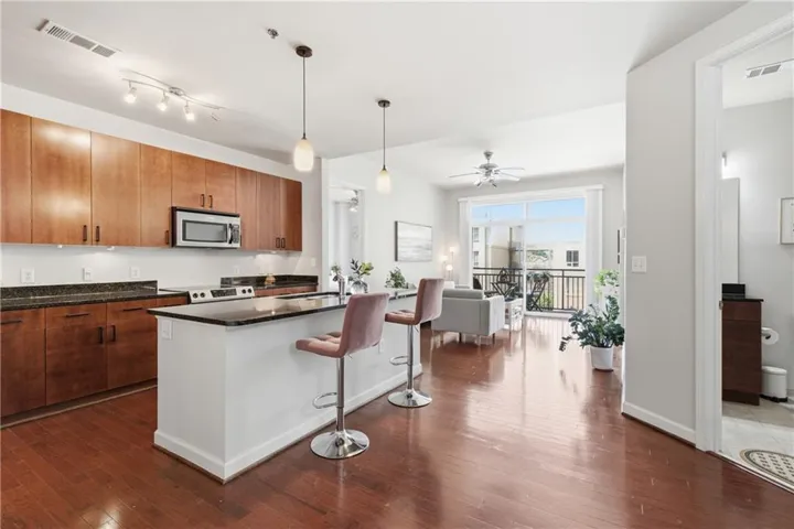 Open-concept living space with rich wood-finish flooring, a kitchen featuring dark countertops and warm wood cabinetry, a breakfast bar island with pendant lighting, a ceiling fan, and large glass doors opening to an outdoor balcony