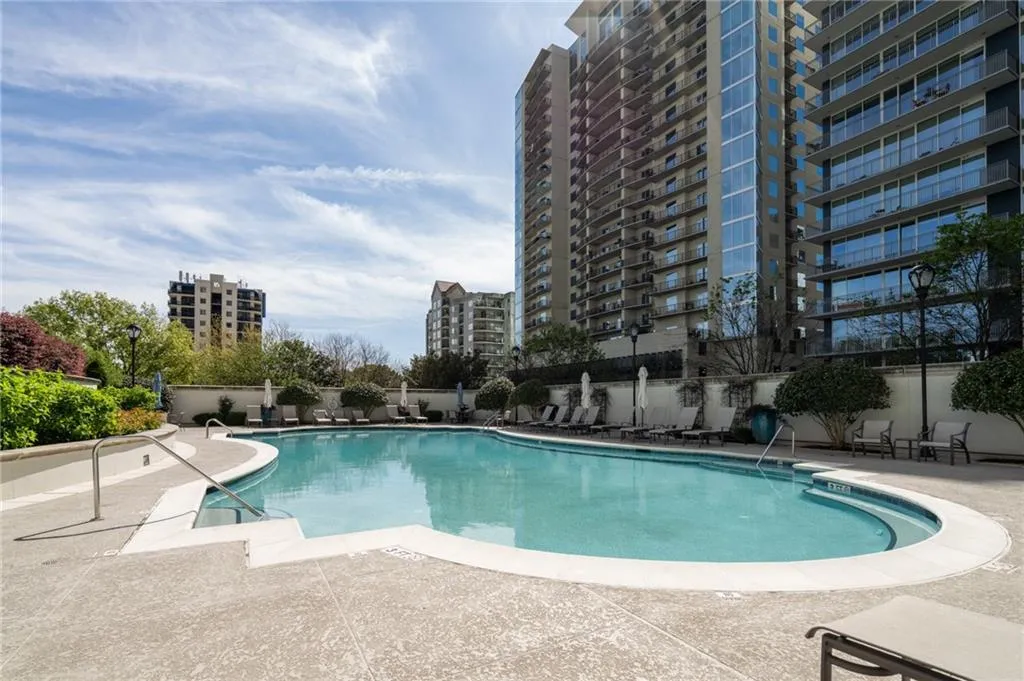 Curved swimming pool with integrated steps, surrounded by a textured concrete deck, manicured hedges, and a privacy wall