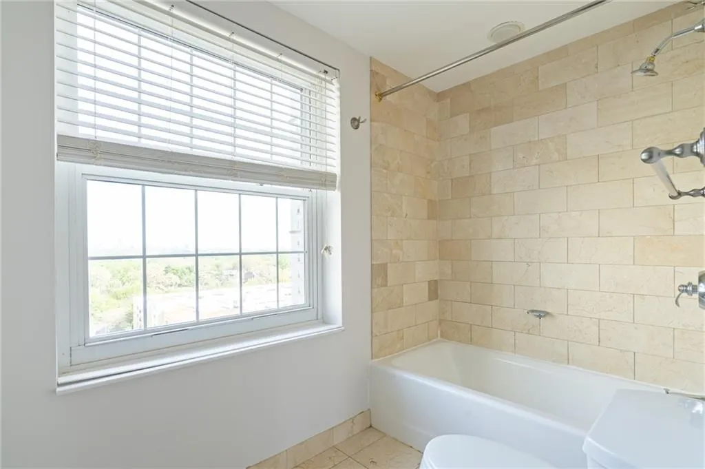White framed window with horizontal blinds, featuring a white tub, light-toned stone tile surround, and chrome fixtures
