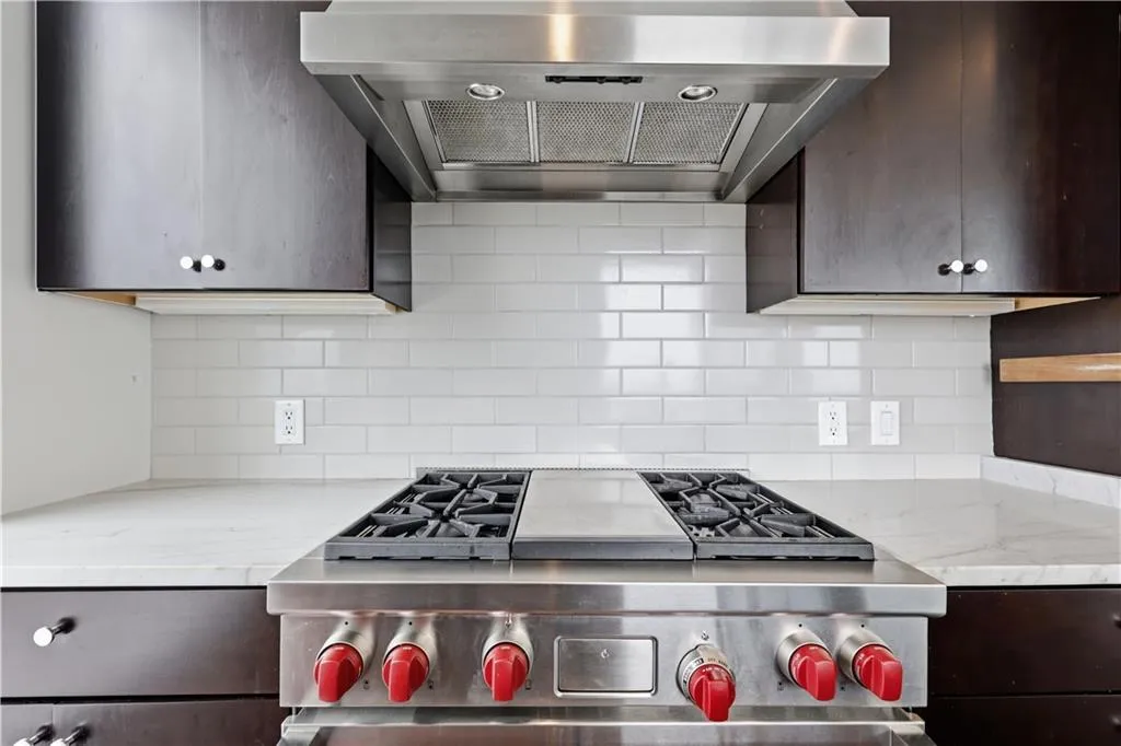 Stainless steel range with a griddle, sleek range hood, subway tile backsplash, quartzite countertop, and dark wood-finish cabinetry