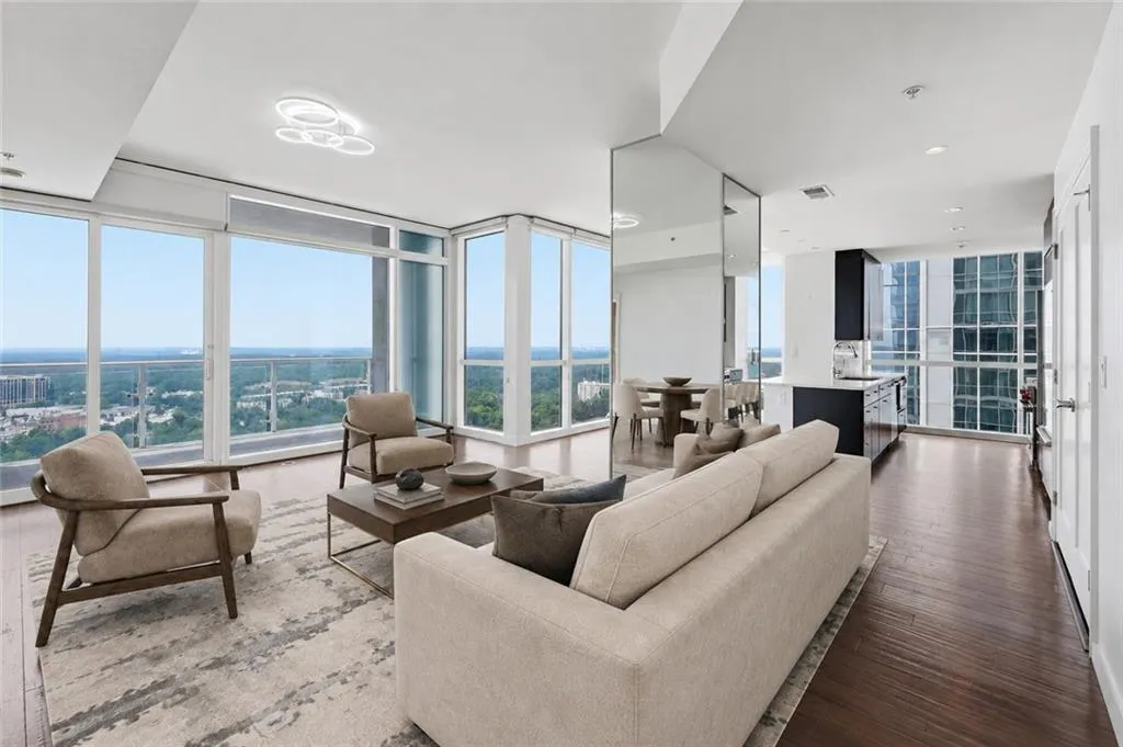VIRTUALLY STAGED-Expansive windows showcasing an elevated panorama, dark wood-finish flooring, a modern circular ceiling light fixture, a mirrored wall feature, and a kitchen with dark cabinetry and a light-colored countertop
