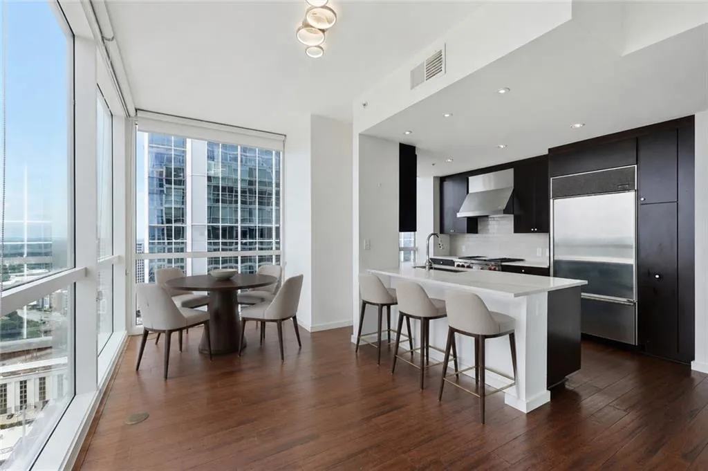 VIRTUALLY STAGED-Open-concept living space with expansive floor-to-ceiling windows, rich wood-finish flooring, and a contemporary kitchen featuring dark cabinetry, an integrated stainless steel refrigerator, and a kitchen island with seating