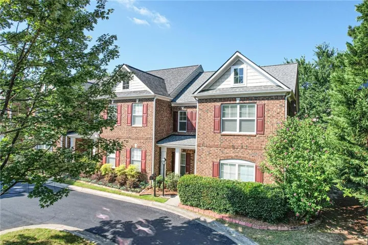 Brick exterior townhome featuring a gabled roofline with dormer windows, red-shuttered windows, and mature landscaping