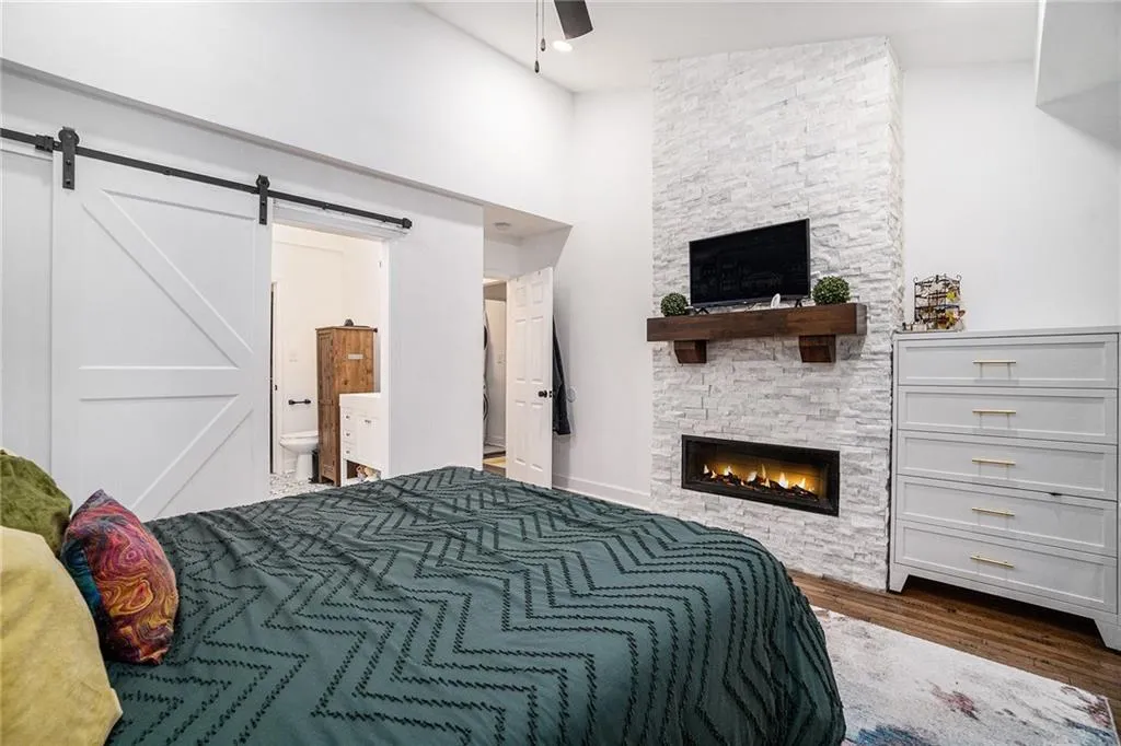 Bedroom with dark wood finished floors, a stone fireplace, a barn door, connected bathroom, and ceiling fan