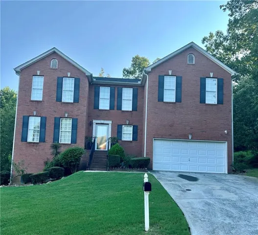 Brick exterior with multiple windows featuring dark shutters, an attached garage with a white door, a paved driveway, and a manicured front lawn
