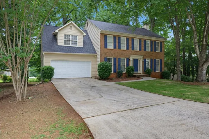 Traditional-style home featuring a brick facade with blue window shutters, an attached two-car garage, a dormer window, a concrete driveway, and mature trees framing the property