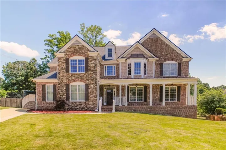 Traditional-style residence featuring a brick and stone facade, covered porch with white columns, gabled rooflines, dark window shutters, and a well-maintained front lawn