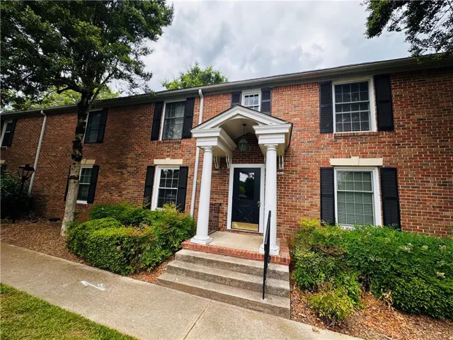 Brick exterior featuring a portico with white columns, a dark entry door with a golden kick plate, and dark shutters on multiple windows
