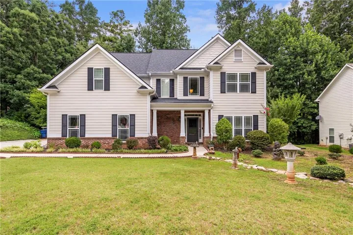 Two-story residence featuring light-colored siding with dark shutters, a front entry with a covered porch, a brick foundation, and a landscaped front yard with mature trees