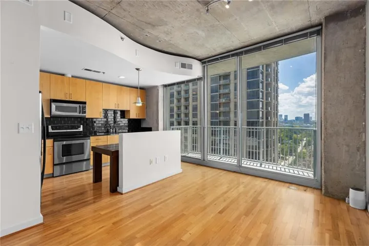 Hardwood flooring throughout, full-height windows with roller shades, exposed concrete ceiling, and a kitchen featuring wood-finish cabinetry, stainless steel appliances, and a dark tile backsplash