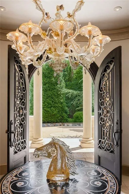 The custom iron and glass entry doors open to a private courtyard framed by columns and lush greenery.