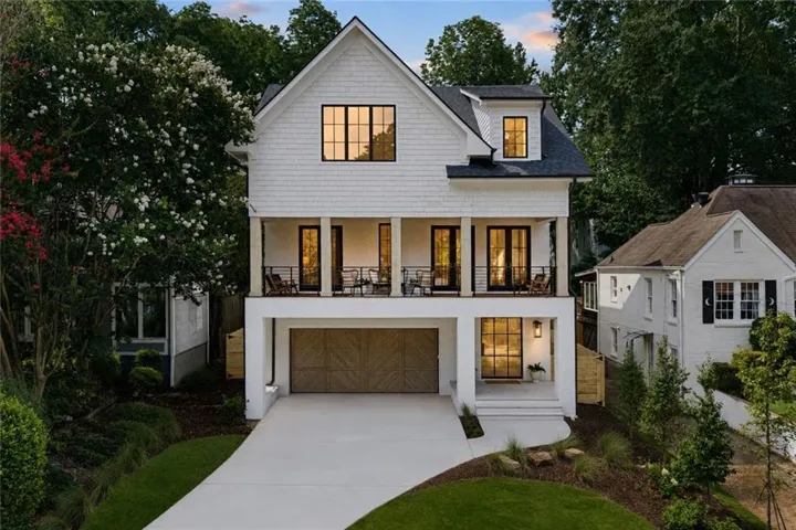 White brick and shingle siding with dark trim windows, covered front entry porch, upper balcony with metal railings, attached garage with wood-finish door, and concrete driveway