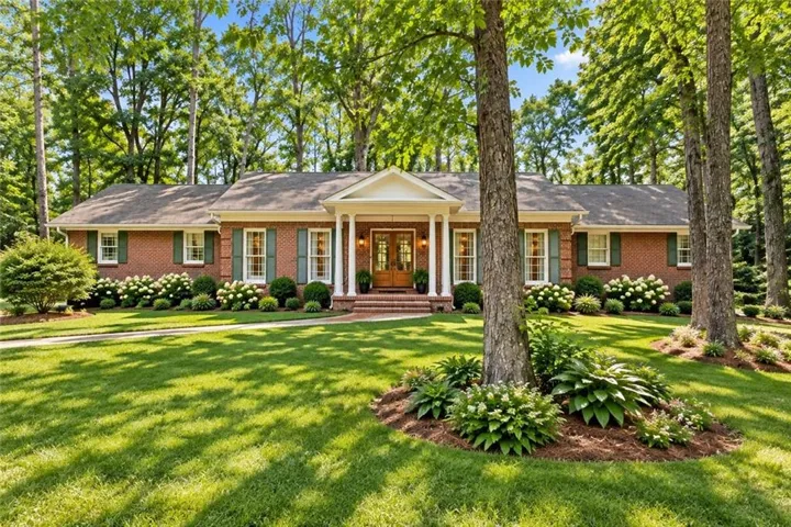 Brick exterior featuring a portico entry with columns, green shutters, and a front lawn with mature trees and shrubbery