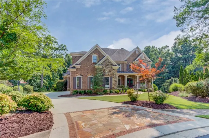 Brick and stone facade, arched windows with shutters, covered front porch, concrete driveway with flagstone inlay, mature landscaping
