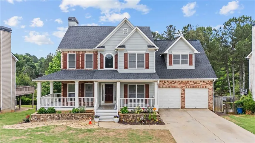 Two-story residence featuring light gray siding, brick accents, a covered front porch with white railing, and a two-car garage with individual doors