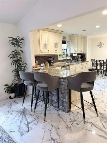 Kitchen featuring cream cabinetry, a stone-finish countertop, mosaic tile backsplash, a kitchen island with stone veneer detailing, and marble-look tile flooring