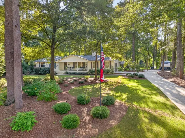 Ranch-style exterior with a covered front porch, mature trees, manicured lawn, established landscaping, and a paved driveway