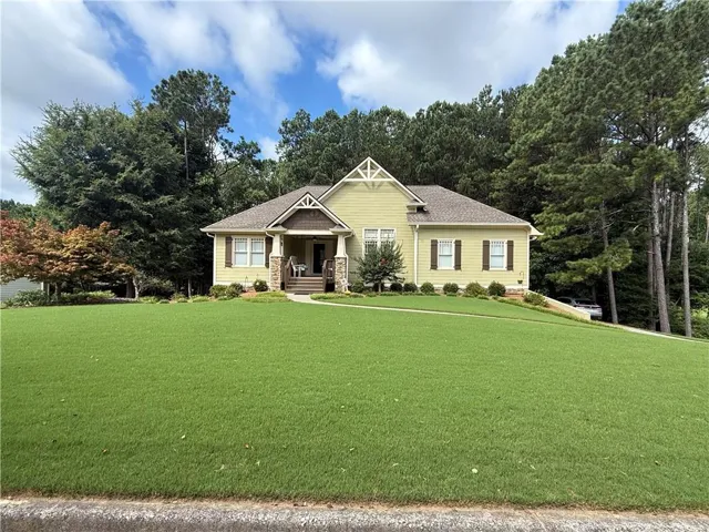 Expansive front lawn with a light yellow siding residence featuring a covered porch with stone columns, dark brown shutters, and a prominent gable with decorative trim