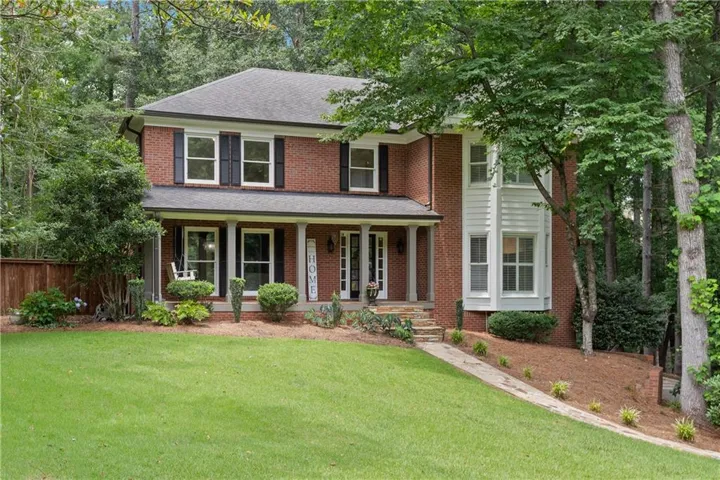 Traditional two-story four-sided brick exterior featuring a covered front porch with white columns, a bay window with white siding, and a gabled roof and professionally landscaped yard
