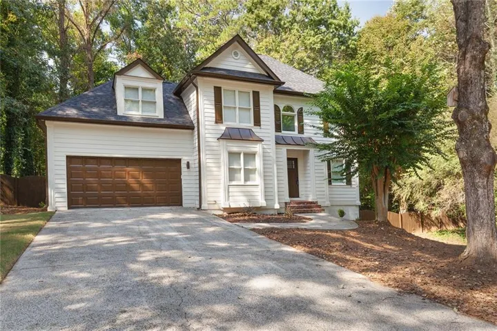 White siding exterior with a dark shingle roof, attached two-car garage, dormer window, front entry porch, and mature trees