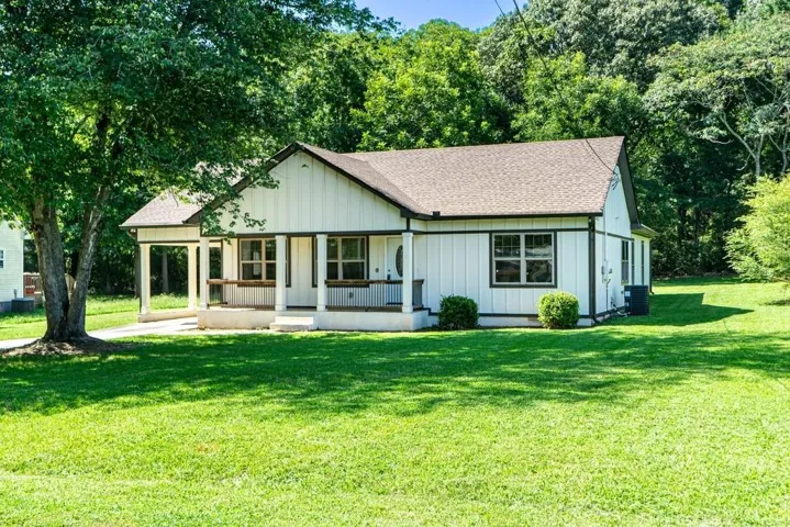 White board-and-batten exterior, gabled roof, covered porch with white columns, concrete porch flooring, and mature tree landscaping