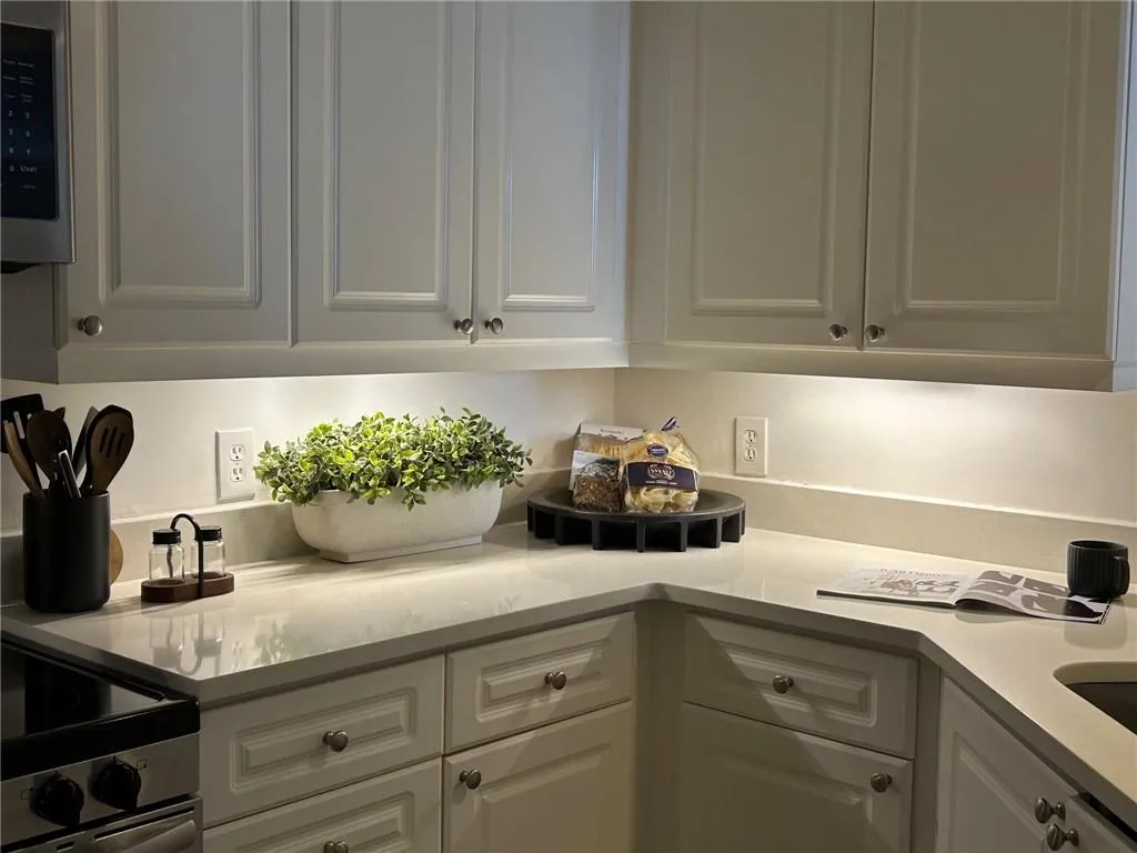 White shaker-style cabinetry with satin nickel hardware, light-toned countertops, under-cabinet lighting, and stainless steel appliances