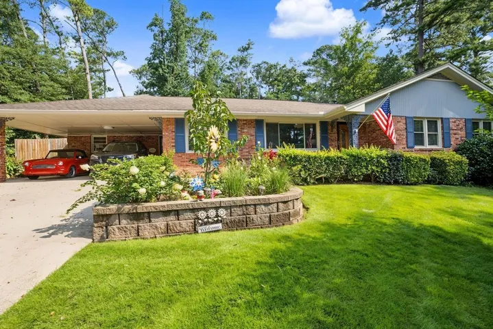 Brick facade with blue shutters, carport, mature trees, and a retaining wall with planting beds