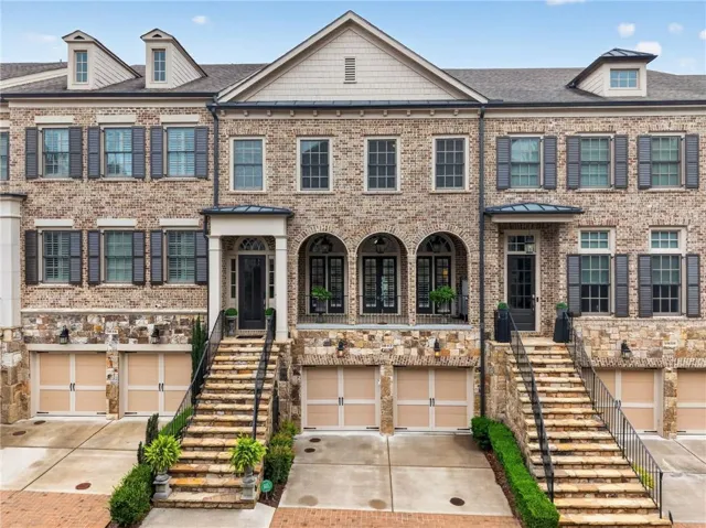 Brick and stone facade townhome featuring dormer windows, dark window shutters, arched entryways, and two-car garage doors