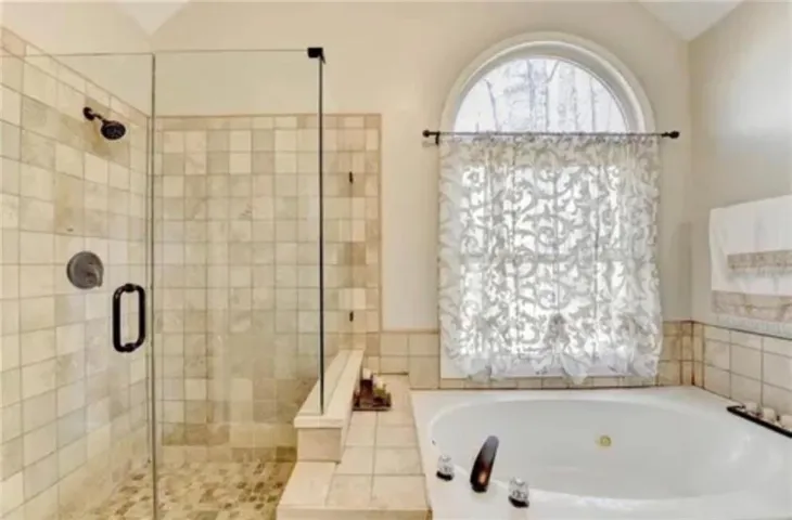 Tile-surrounded soaking tub with an arched window, separate glass-enclosed shower, and tiled walls