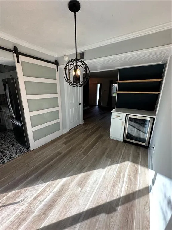 Unfurnished dining area featuring a barn door, light wood-style floors, beverage cooler, crown molding, and a chandelier