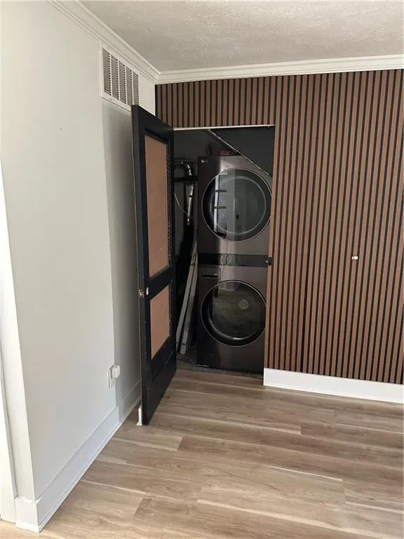 Washroom featuring ornamental molding, a textured ceiling, light wood-style flooring, stacked washer / drying machine, and wallpapered walls