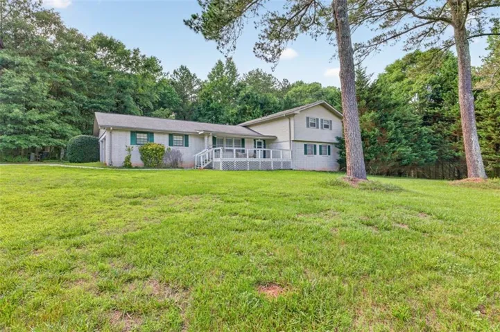 Spacious lawn with mature trees, a home featuring light-colored siding, a front deck with white railings, and green window shutters