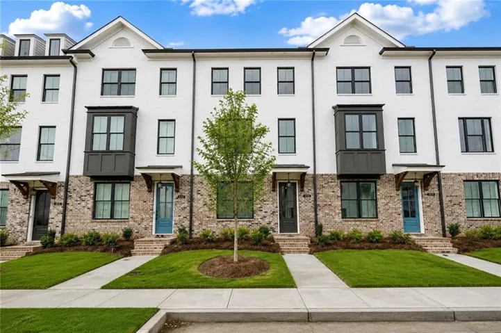 Modern townhome exterior featuring white brick, a dark gray bay window, prominent gables, dark framed windows, and a manicured lawn
