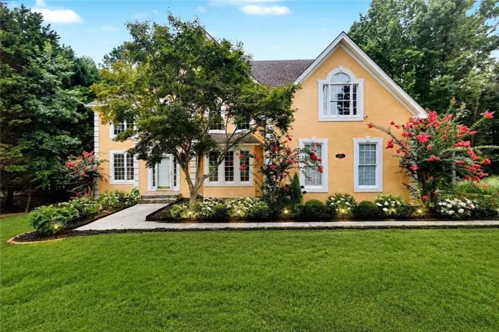 Yellow stucco exterior with white trim, arched window, lush landscaping, mature trees, and a concrete pathway
