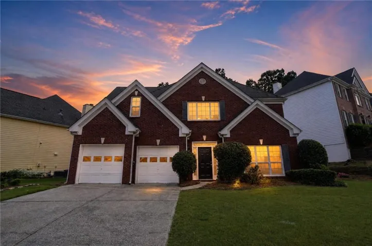 Brick Colonial home featuring a two-car garage with paneled doors, illuminated front entry with arched transom window, and manicured lawn with mature landscaping