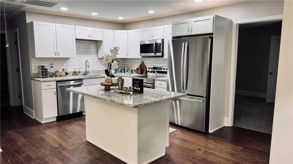 Kitchen featuring appliances with stainless steel finishes, a kitchen island, light stone countertops, decorative backsplash, and dark wood-type flooring