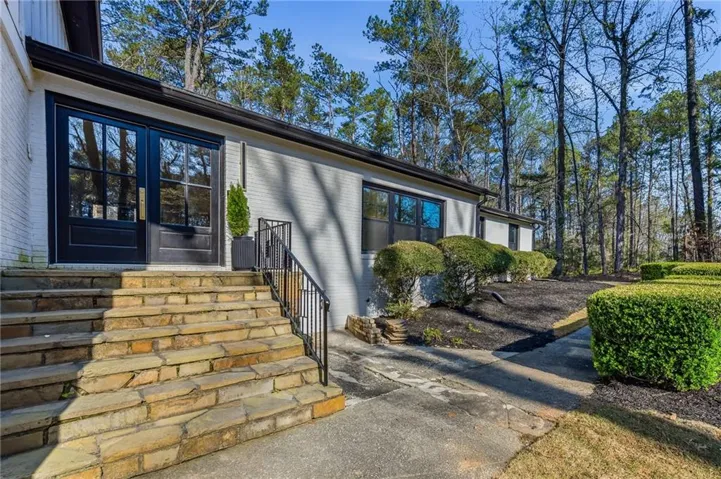 Modern residence showcasing a striking black-framed facade with expansive glass panels, wide stone-clad entry steps with metal railings, and manicured landscaping including sculpted evergreen hedges and mature pine trees throughout the grounds