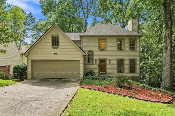 Two-story residence featuring light-colored siding, an attached two-car garage, multiple windows, a prominent chimney, and mature trees surrounding the property