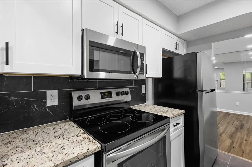 Kitchen close-up featuring stainless steel appliances, light stone countertops, Dark tile backsplash, & white cabinetry.